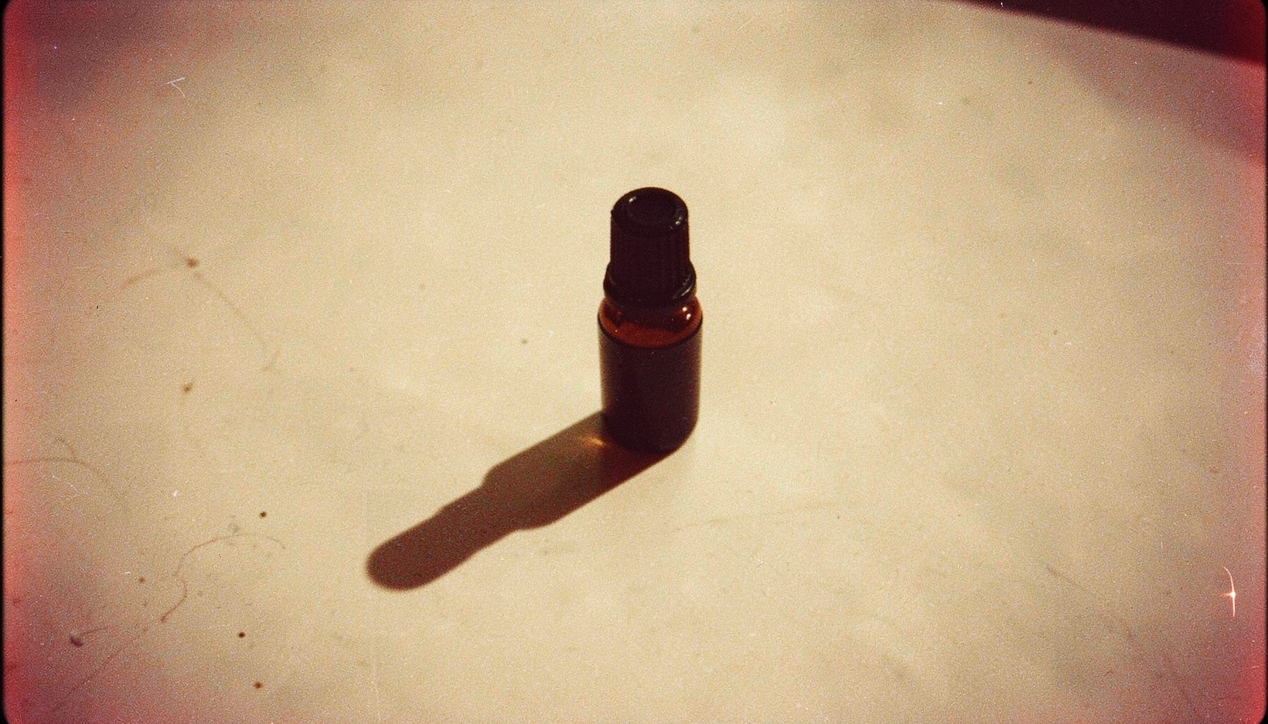 Analog film still of a single dropper bottle casting a long shadow across a pale surface, subdued beige and brown palette