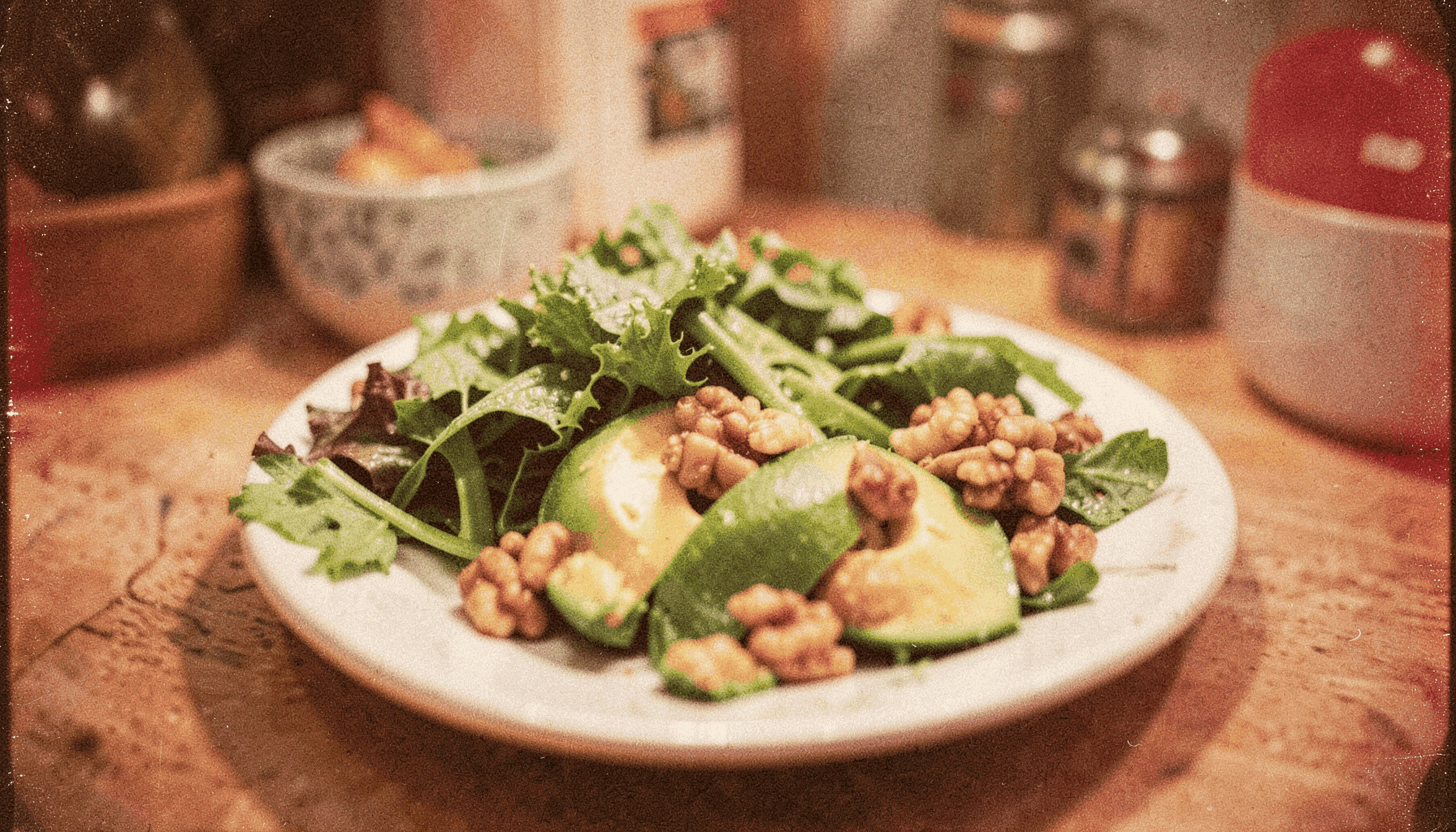 Analog film photograph of a plate with fresh leafy greens, avocado, and nuts, warm kitchen lighting with visible grain texture